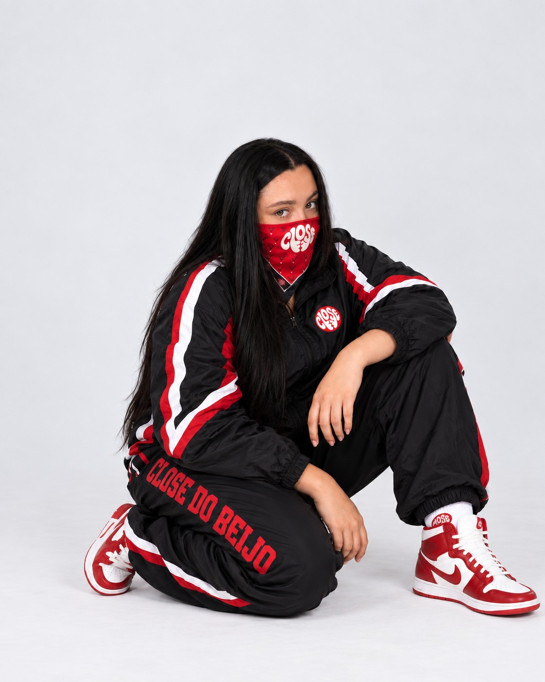 Mya Jokeman, de bandana vermelha cobrindo o rosto e agasalho preto com a marca Close do Beijo, agachada